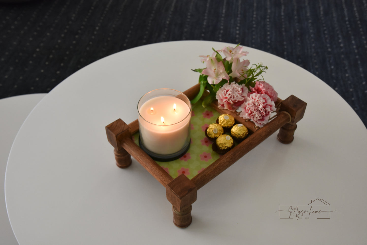 Decorative setup with a lit candle, flowers, and chocolates on a wooden khaat platter.