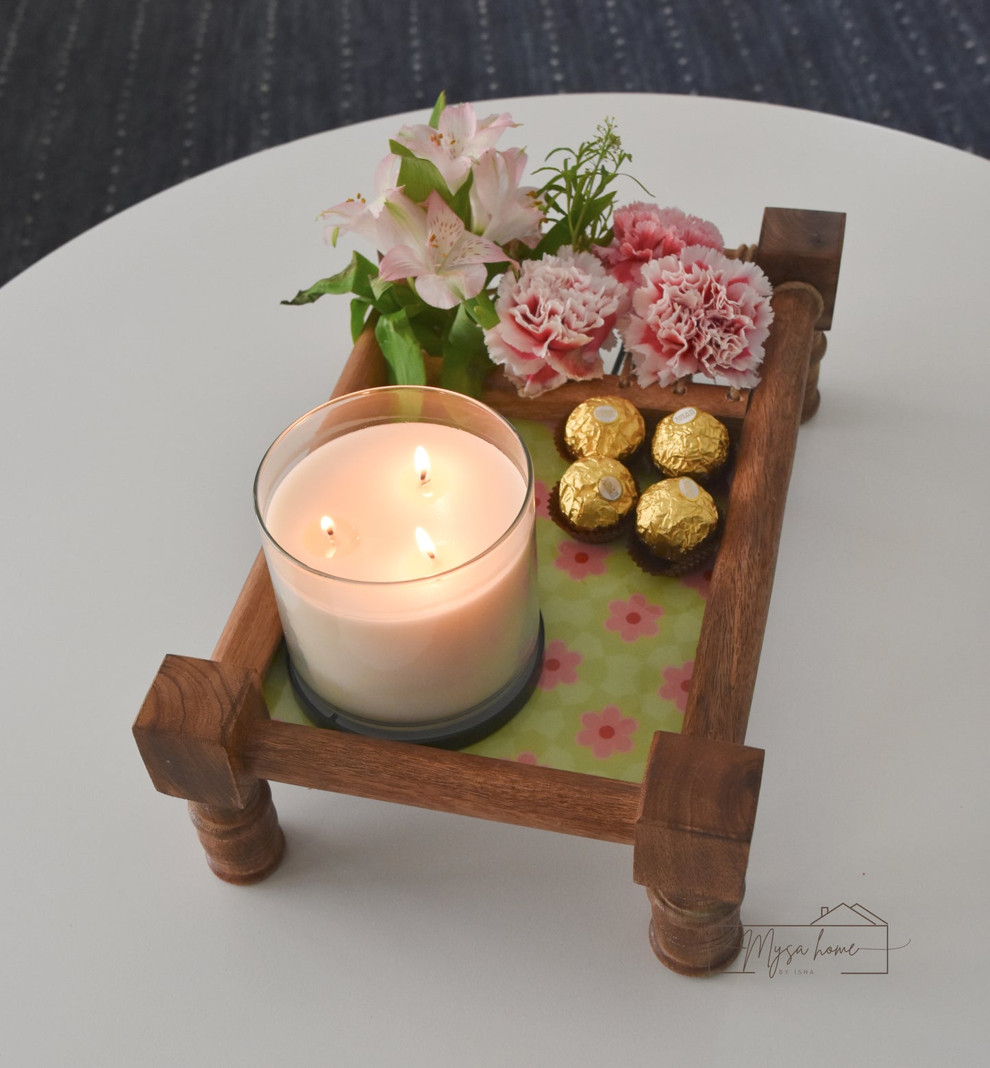 Decorative wooden tray with a candle, flowers, and chocolates on a white surface.
