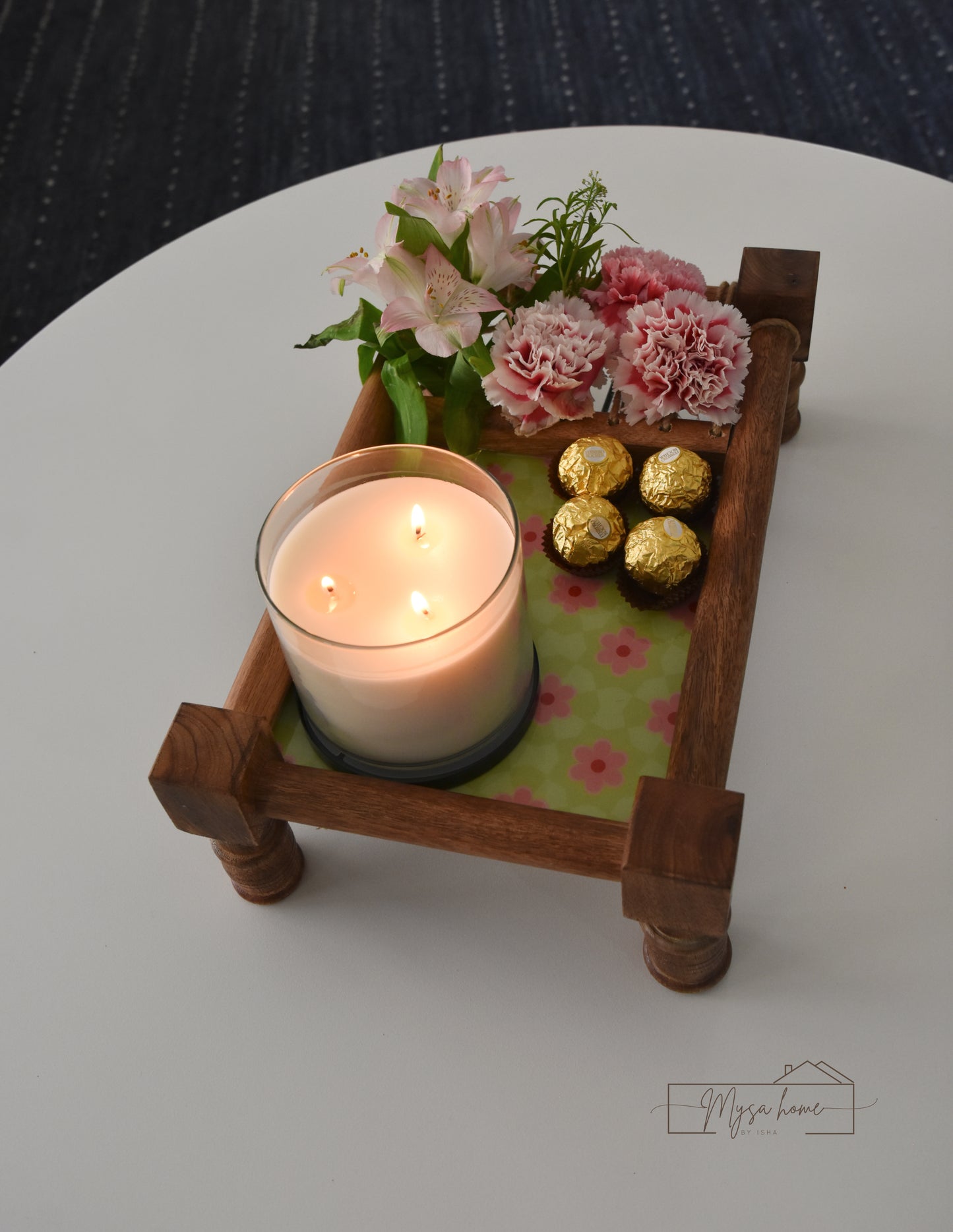 Wooden tray with a lit candle, flowers, and chocolate on a white surface.