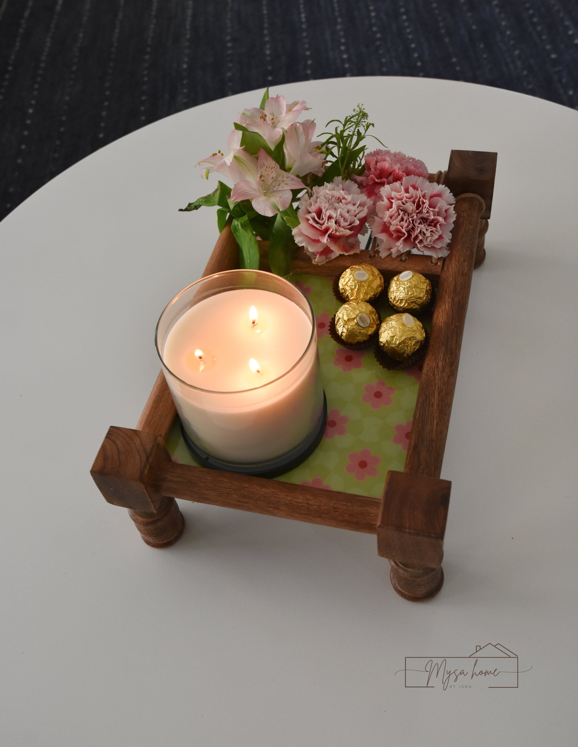 Wooden tray with a lit candle, flowers, and chocolate on a white surface.